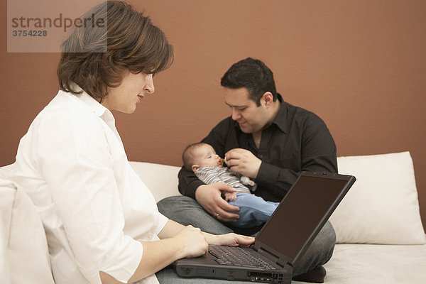 Junge Frau arbeitet am Computer während ihr Mann sich um das Baby kümmert