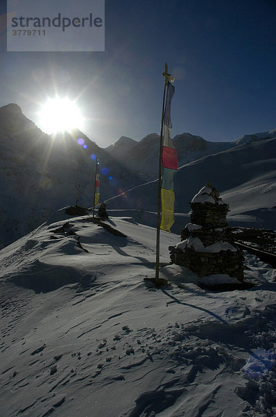 Helle Strahlen der Morgensonne an Gebetsfahne und Chörten im verschneiten Hochgebirge nahe Thorung La Pass Annapurna Region Nepal