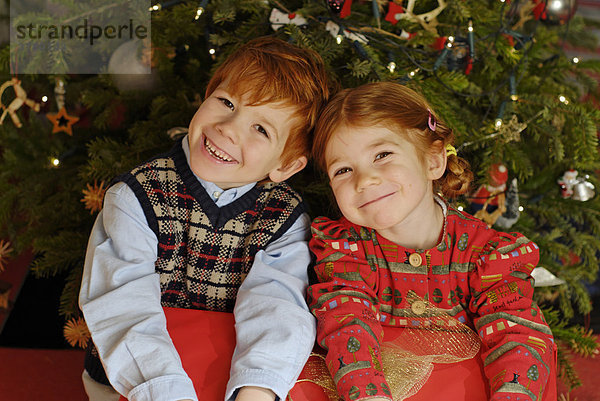 Zwei Kinder sitzen mit einem Geschenk unter dem Weihnachtbaum