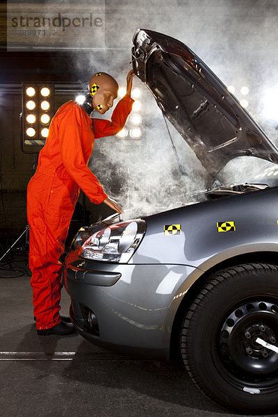 Ein Crashtest-Dummy  der unter der Motorhaube eines rauchenden Autos prüft.