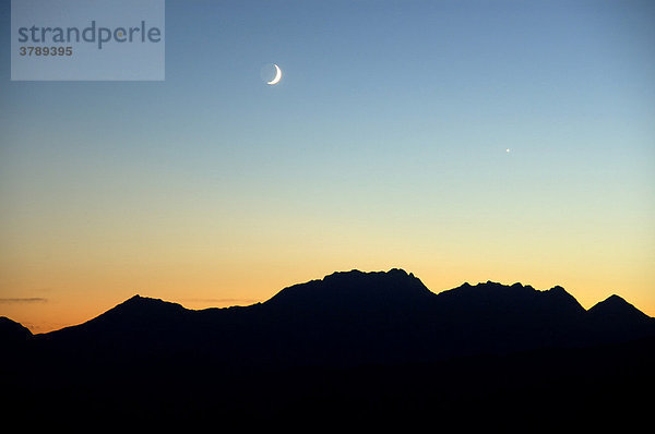 Abendhimmel mit Mond und Abendstern über Bergkette vor orangenem Himmel Hochsavoyen Haute-Savoie Frankreich