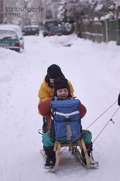 Zwei Schulkinder auf Schlitten werden auf verschneiter Straße gezogen