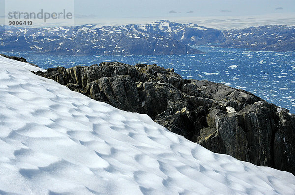 Weiter Blick über Schneefeld und Felsen auf Eisberge im Sermilik Fjord Ostgrönland