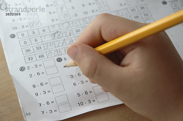Hausaufgaben: Hand eines Kindes mit gelbem Bleistift  im Hintergrund ein Blatt mit Rechenaufgaben