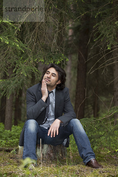Junger Mann sitzt nachdenklich im Wald auf einem Baumstumpf