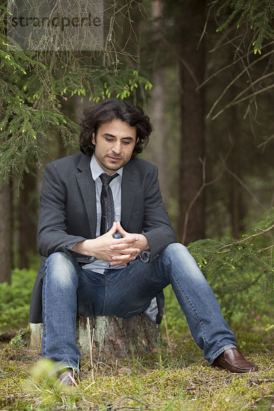 Junger Mann sitzt nachdenklich im Wald auf einem Baumstumpf