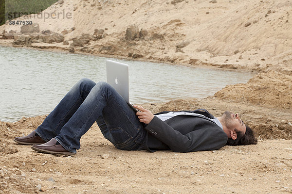 Junger Mann liegt mit Laptop am Seestrand und denkt nach