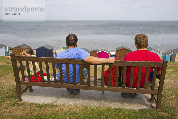Familie auf einer Bank mit Blick auf Strandhütten und Meer bei Tankerton in der Nähe von Whitstable  Kent  England  Vereinigtes Königreich  Europa