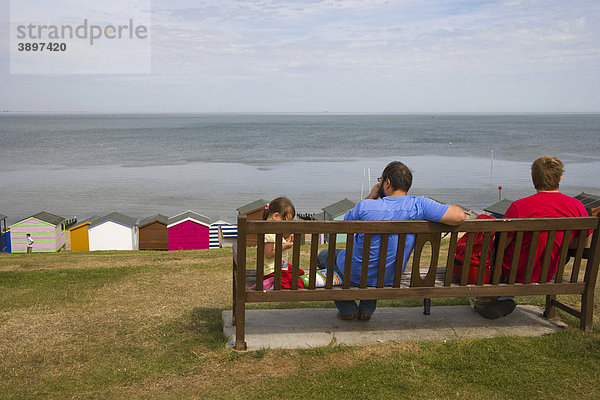 Familie auf einer Bank mit Blick auf Strandhütten und Meer bei Tankerton in der Nähe von Whitstable  Kent  England  Vereinigtes Königreich  Europa