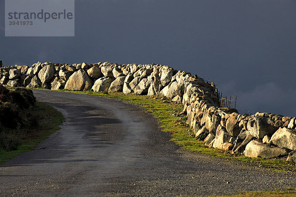 Sonnenbeschienener  steinerner Begrenzungszaun und Straße mit dunkle Gewitterwolke  Republik Irland  Europa
