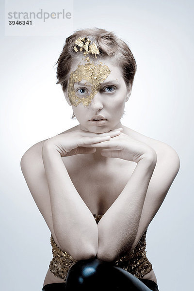 Gesicht von junger Frau mit Blattgold Makeup und Paillettentop auf Arme gestützt