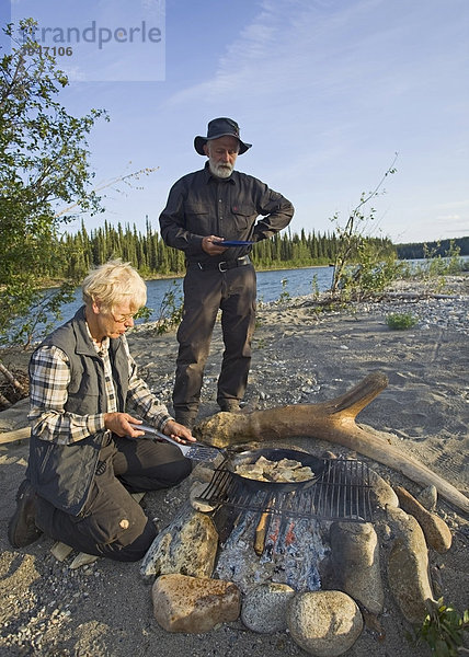 Eine Frau brät Fischfilets in einer Pfanne auf dem Lagerfeuer  ein Mann mit Teller wartet daneben  oberer Liard River  Yukon Territory  Kanada