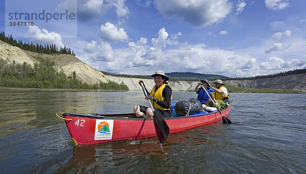 Eine Familie mit Sohn paddelt in einem Kanu auf dem Teslin River  dahinter die hohe Böschung des Flussufers  Yukon Territory  Kanada