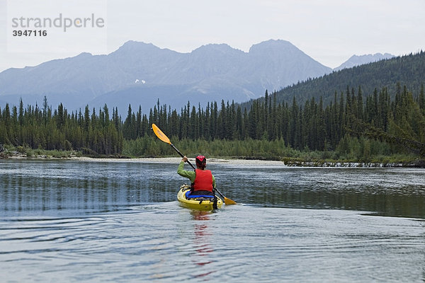 Eine Frau paddelt in einem Kajak auf dem oberen Liard River  dahinter die Pelly Mountains  Yukon Territory  Kanada