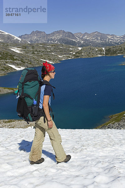 Junge Wanderin mit Rucksack wandert über ein Schneefeld  Panorama beim Gipfel des historischen Chilkoot Passes  Chilkoot Trail  Crater Lake See dahinter  alpine Tundra  Yukon Territory  British Columbia  BC  Kanada