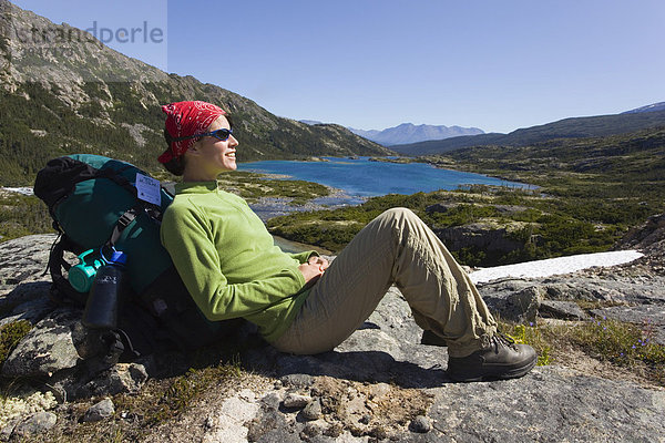 Junge Wanderin  Rucksacktourist  auf einem Felsen sitzend  ruht aus und genießt das Panorama  Deep Lake See  historischer Chilkoot Pass  Chilkoot Trail Wanderweg  Yukon Territory  British Columbia  BC  Kanada