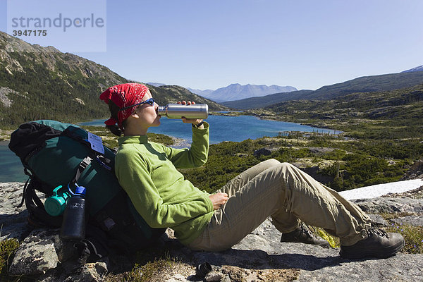 Junge Wanderin  Rucksacktourist  auf einem Felsen sitzend  trinkt  ruht aus und genießt das Panorama  Deep Lake See  historischer Chilkoot Pass  Chilkoot Trail Wanderweg  Yukon Territory  British Columbia  BC  Kanada