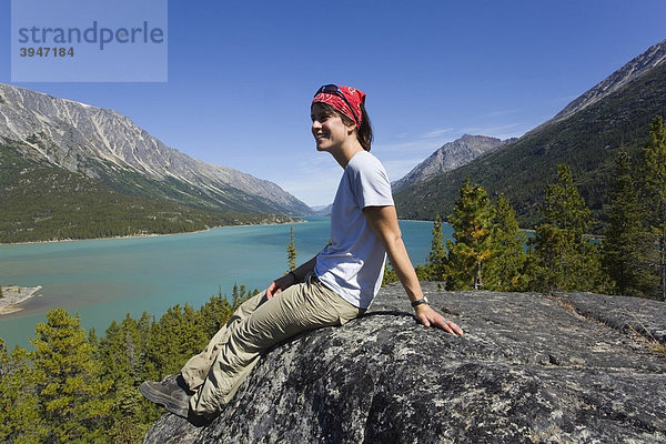Junge Wanderin  Rucksacktourist  auf einem Felsen sitzend  ruht aus und genießt die Aussicht auf den Lake Bennett See  historischer Chilkoot Pass  Chilkoot Trail Wanderweg  Yukon Territory  British Columbia  BC  Kanada