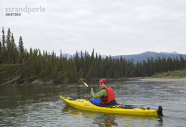 Junge Frau paddelt in einem Kajak  Kajakfahren  oberer Liard River  dahinter die Pelly Mountains  Yukon Territory  Kanada