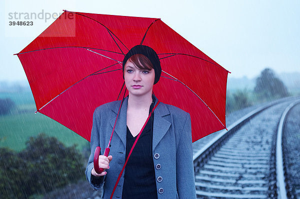 Junge Frau mit einem roten Regenschirm geht auf Bahngleisen