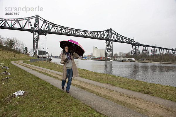 Junge Frau  30-35 Jahre  mit buntem Regenschirm geht an einem nasskalten Wintertag am Nord-Ostsee-Kanal spazieren  Schleswig-Holstein  Deutschland  Europa