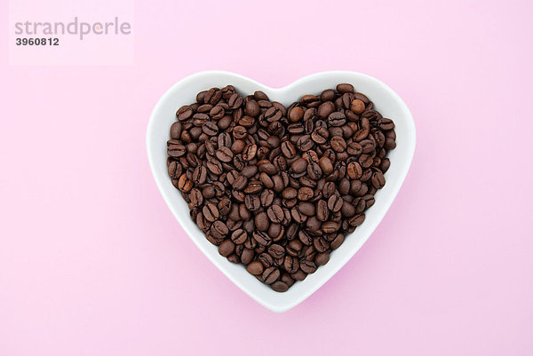 Herzförmige weiße Schale gefüllt mit Kaffeebohnen auf Rosa