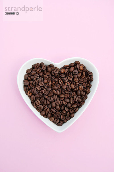 Herzförmige weiße Schale gefüllt mit Kaffeebohnen auf Rosa