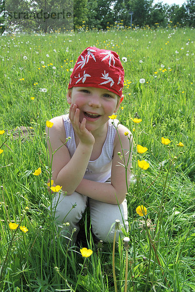 Glückliches Mädchen  5 Jahre  mit rotem Kopftuch auf einer Sommerwiese mit Sumpfdotterblumen und Pusteblumen