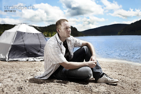 Junger Mann sitzt vor seinem Zelt am See und blickt dabei in die Ferne