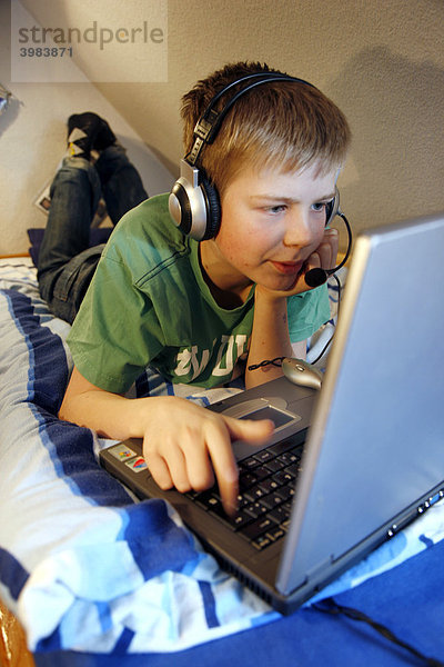 Junge  13 Jahre alt  arbeitet mit seinem Computer zuhause in seinem Kinderzimmer auf dem Bett  surft im Internet auf einer Chat-Seite  spricht mit anderen Chat-Teilnehmern über sein Headset