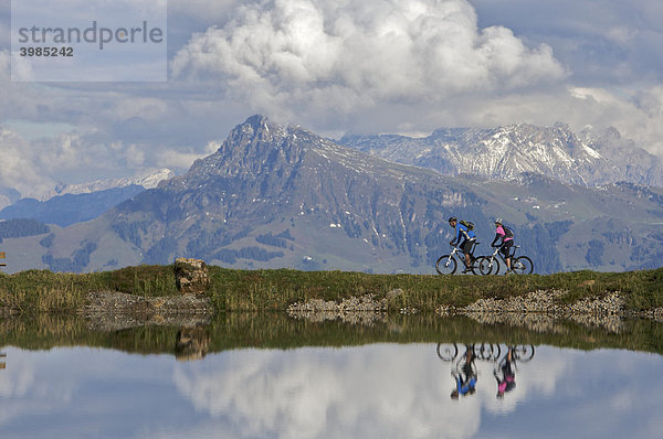 Mountainbike-Fahrerin und -Fahrer spiegeln sich im Wasserreservoir Salvensee an der Hohen Salve  dahinter Kitzbüheler Horn  Tirol  Österreich