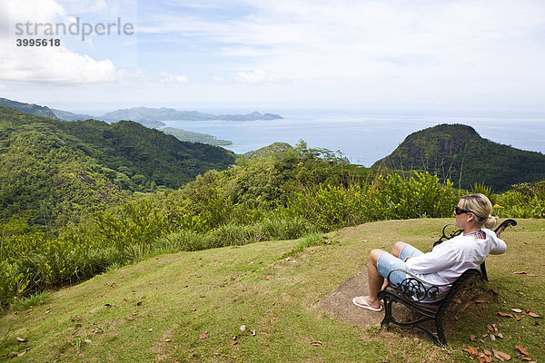 Frau sitzt auf einer Bank im Nationalpark Morone  hinten der Strand von Grand Anse Insel Mahe  Seychellen  Indischer Ozean  Afrika