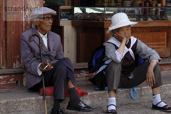 Alter Mann mit Stock  Hut und Brille und alte Frau mit Sonnenhut und Tracht der Naxi Minderheit sitzen vor einem Geschäft in China  Lijiang  Yunnan  China