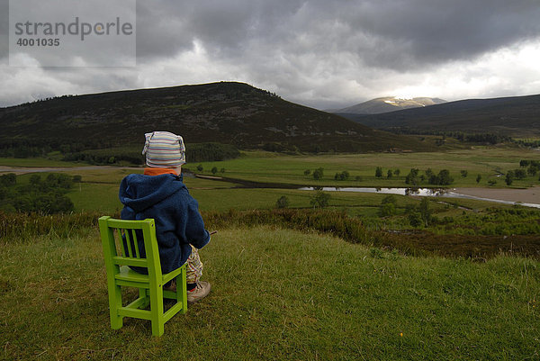 Kleines Mädchen sitzt auf einem grünen Kinderstuhl vor der Landschaft der schottischen Highlands mit Fluss und Wäldern  Schottland  Europa