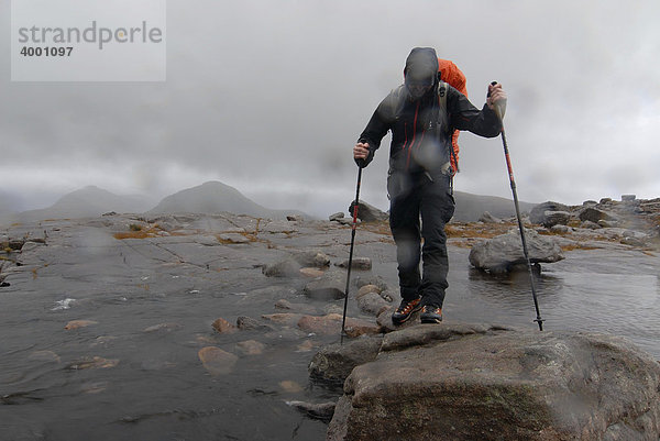 Wanderer mit Teleskopstöcken und Regenanzug beim Überqueren eines Flusses im schottischen Regen  schottische Highlands  Liathach  Torridon Schottland  Großbritannien  Europa