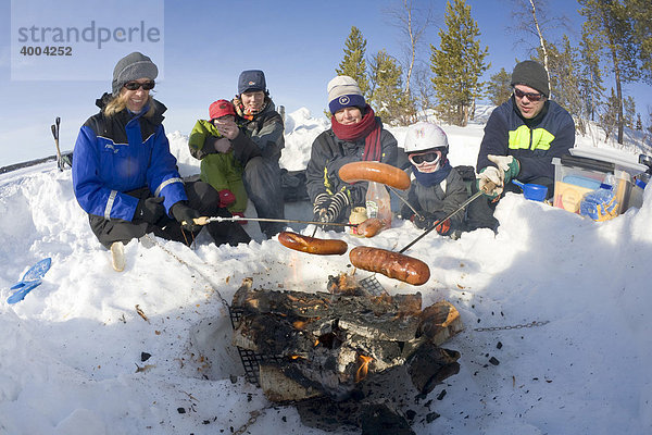 Drei Frauen und ein Mann in den Vierzigern grillen zusammen mit zwei Kindern im Schnee ihre Würste über einem offenen Feuer in Kiruna  Lappland  Nord-Schweden  Schweden