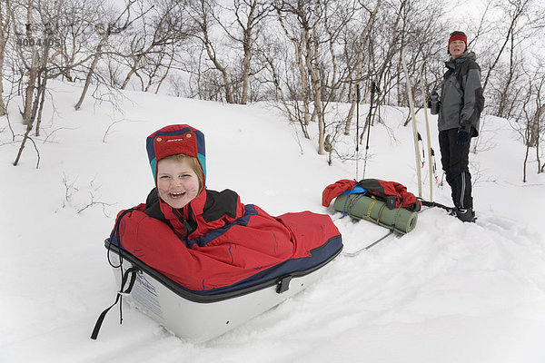 Schlittenfahrt in Lappland  vierjähriges Mädchen sitzt in einer Pulka  Mutter steht dahinter  Kiruna  Lappland  Nord-Schweden  Schweden