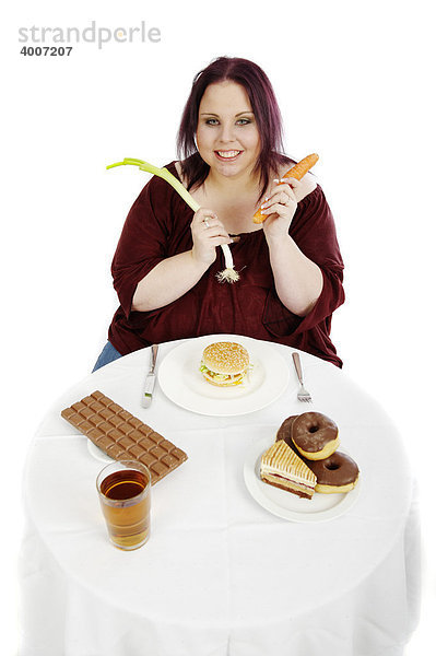 Junge dicke Frau mit Gemüse vor Tisch mit Hamburger und Torten