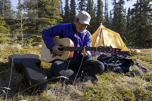 Asiat sitzt im Gras und spielt Gitarre  dahinter ein Zelt  Camping  Yukon Territory  Kanada  Nordamerika