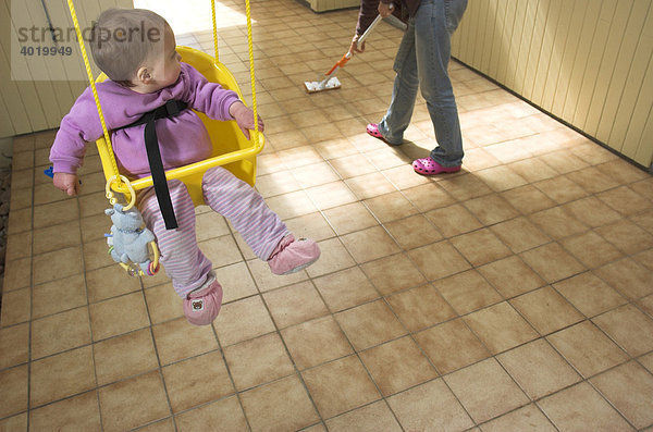 Baby  Mädchen  in einer Schaukel  nach hinten zu ihrer Mutter gedreht  die den Kachelboden wischt