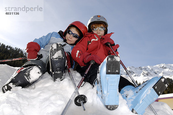 Zwei Kinder mit Skischuhen Skihelm und Sonnenbrillen sitzen auf einem Schneehügel