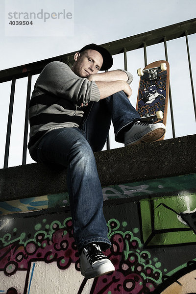 Junger Mann sitzt mit seinem Skateboard auf einer mit Graffiti besprühten Wand