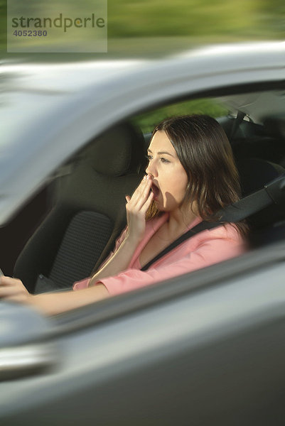 Junge Frau sitzt müde und gähnend am Steuer ihres Autos