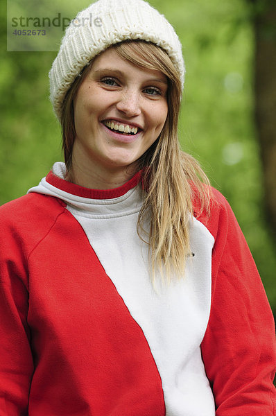 Portrait junge  schöne  sportliche  blonde Frau im Sommer im Park  lächelt  trägt Mütze und roten Kapuzenpullover
