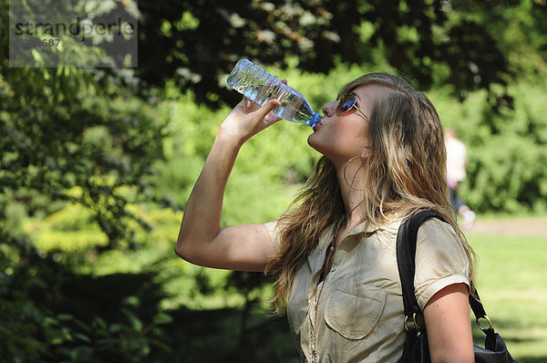 Blonde  langhaarige Frau mit Sonnenbrille und beigem Kleid trinkt Wasser aus Plastikflasche im Park