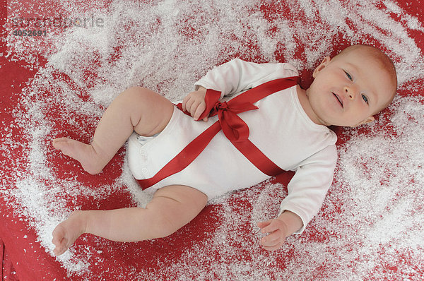 Weihnachts-Baby  3 Monate  liegt mit weißem Body und roter Schleife auf rotem Untergrund mit Schnee  Kunstschnee und lacht