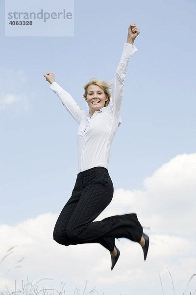 Junge blonde Frau in Business-Kleidung springt vor blauem Himmel und reißt die Arme hoch