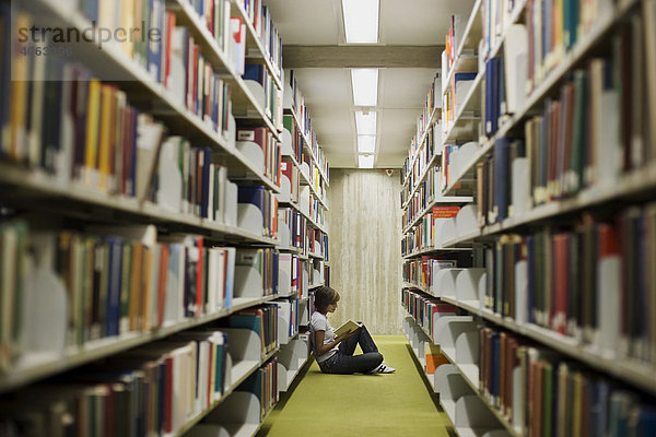 Junge dunkelhaarige Frau  Studentin  sitzt zwischen den Bücherregalen in der Bibliothek und liest in einem Buch