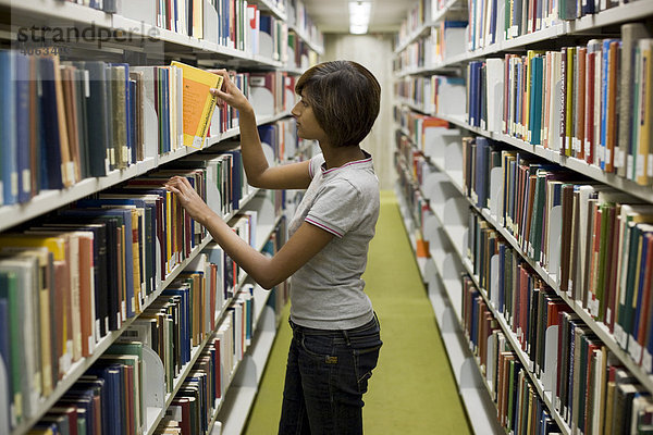 Junge dunkelhaarige Frau  Studentin  greift nach einem Buch aus einem Bücherregal in der Bibliothek