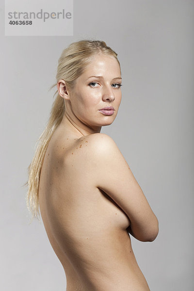 Junge blonde Frau steht mit unbekleidetem Oberkörper vor Grau und verdeckt mit ihren Armen die Brust